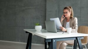 Woman using phone and laptop in office while reading papers